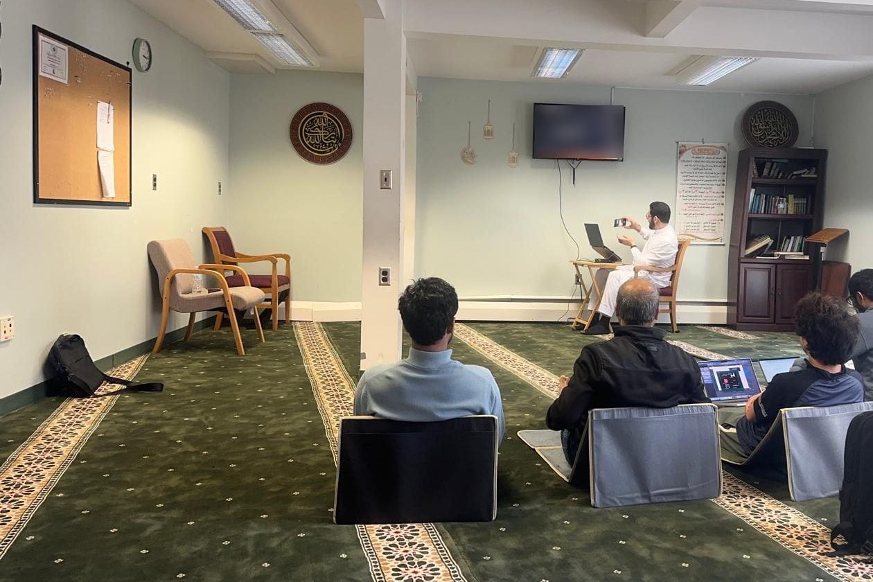 Attendees sit on floor seats in the masjid prayer hall, some with laptops open, facing a speaker at the front.