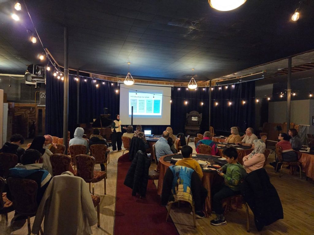 People at round tables watch a presenter beside an Introduction to Islam slide, under strings of lights.