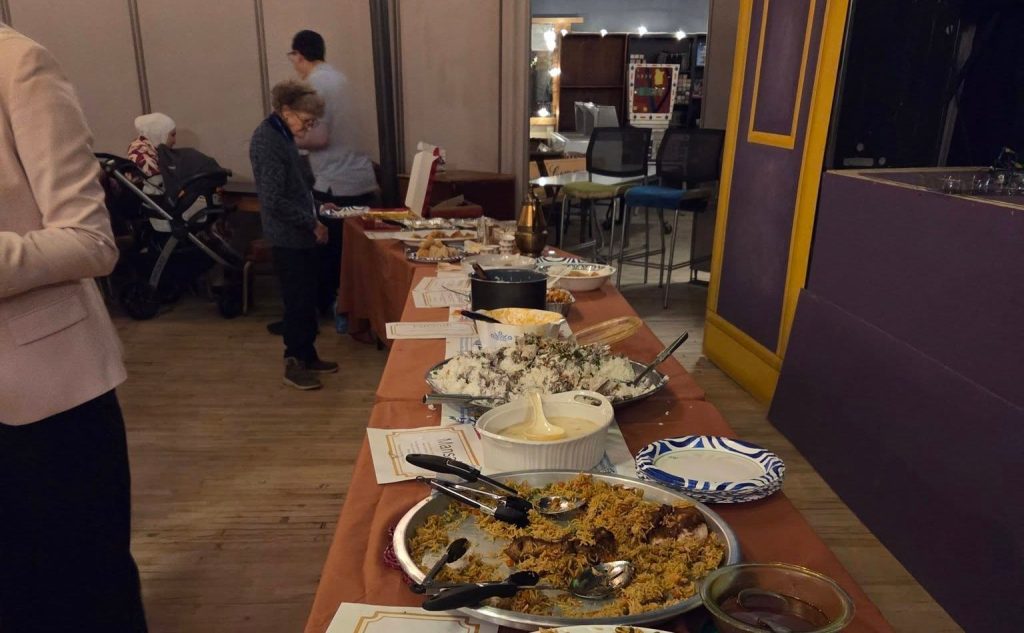 A long buffet table of rice platters and other dishes, with people serving themselves at the far end.