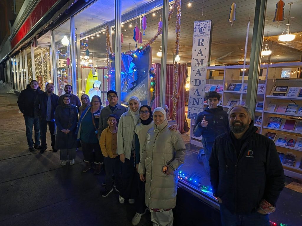 A group of people smiles on the sidewalk outside the decorated Storytelling Center window at night.