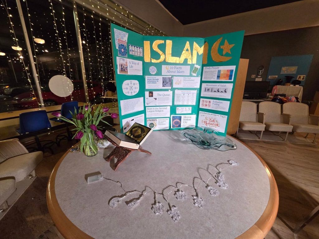 A display board titled Islam stands on a round table with a Quran on a wooden stand and purple tulips.