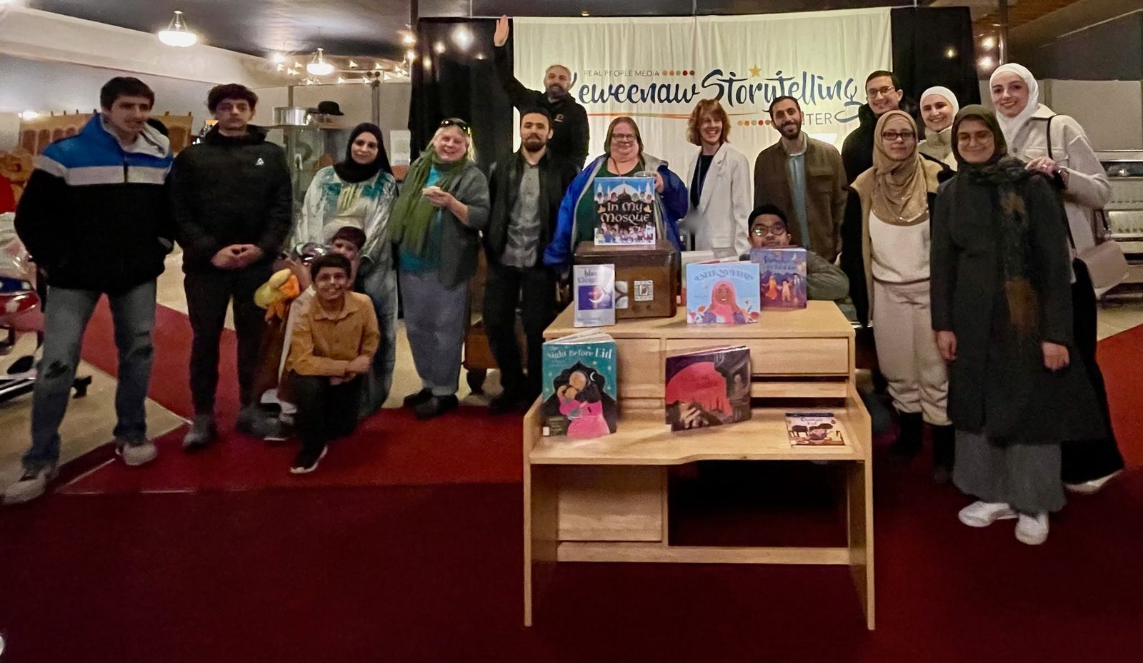 A group poses under the Keweenaw Storytelling Center banner around a small desk of books about Islam, Ramadan and Eid.