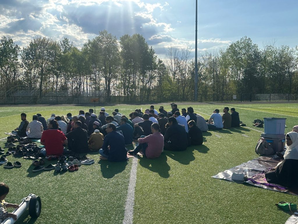 The congregation sits on the field facing a lectern, seen from behind in bright sunlight.
