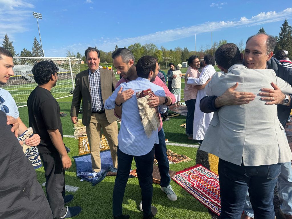 Two pairs of men embrace on the field after the Eid prayer, one holding a prayer rug.