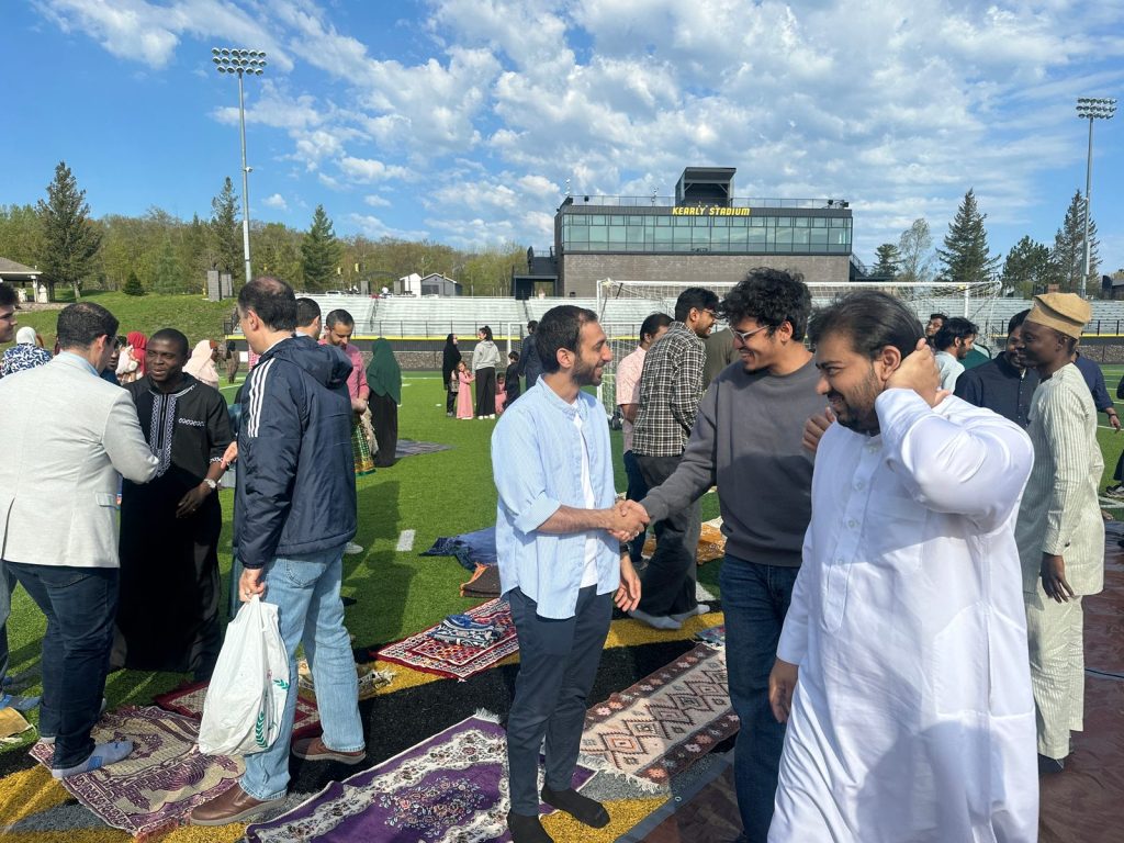 Two young men shake hands on the prayer rugs, with Kearly Stadium behind them.