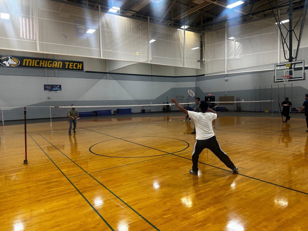 A player in a white shirt swings at the shuttlecock under a Michigan Tech banner.