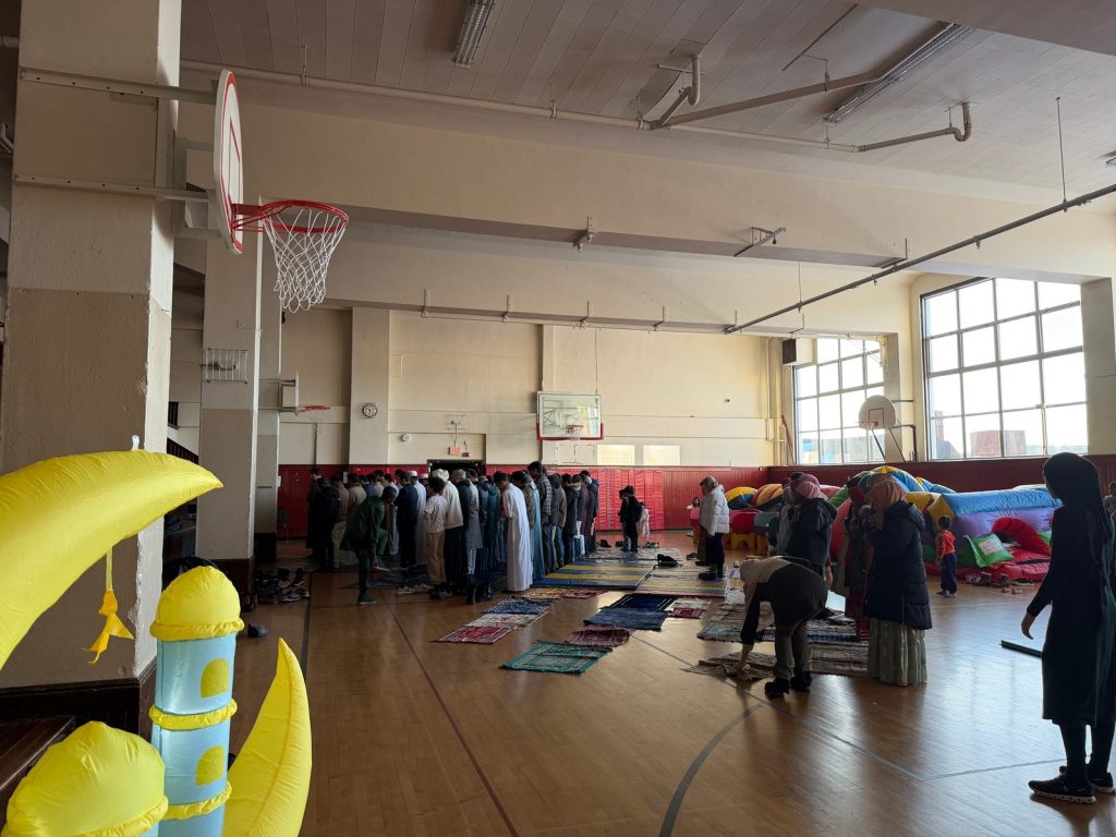 Worshippers stand in rows for the Eid prayer in a gym, with an inflatable crescent decoration in the foreground.