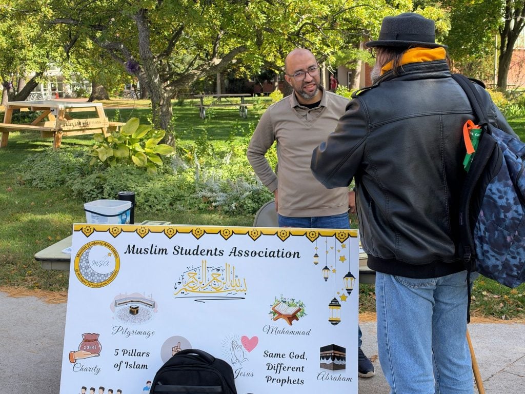 A man at the MSA table talks with a visitor in a leather jacket and hat beside the MSA display board.