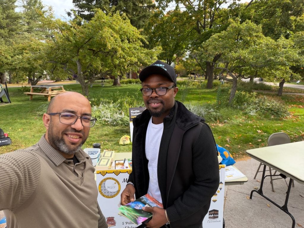 A man from the MSA table and a visitor holding booklets smile for a selfie in front of the table.