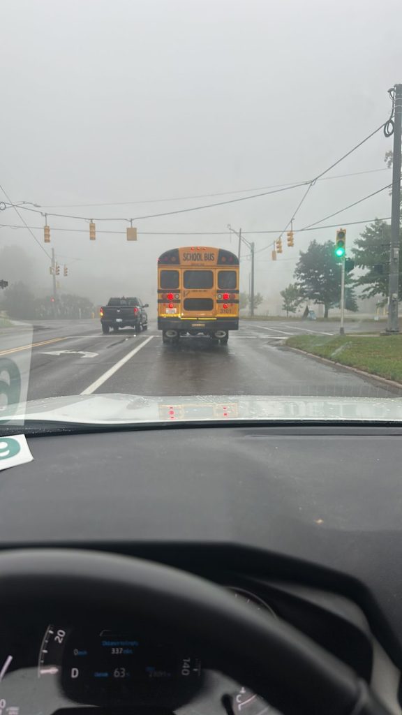 A yellow school bus drives ahead through thick fog at a town intersection, seen through a car windshield.