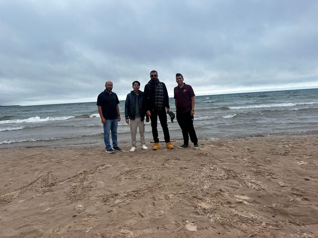 Four men stand together at the water's edge on a sandy beach, with choppy waves and grey clouds behind.