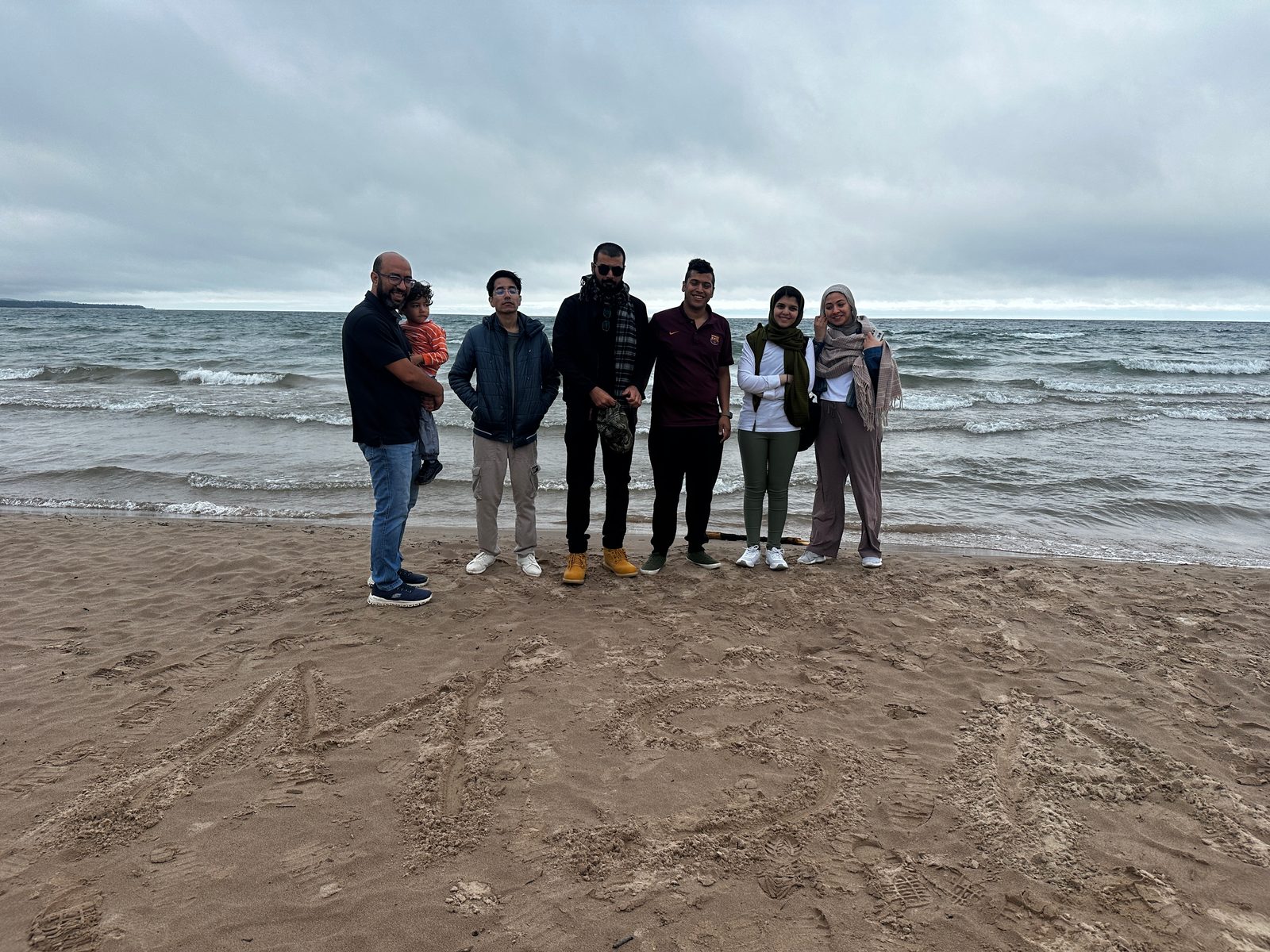 Six adults and a toddler stand together on a windy beach under grey skies, with MSA written in the sand.