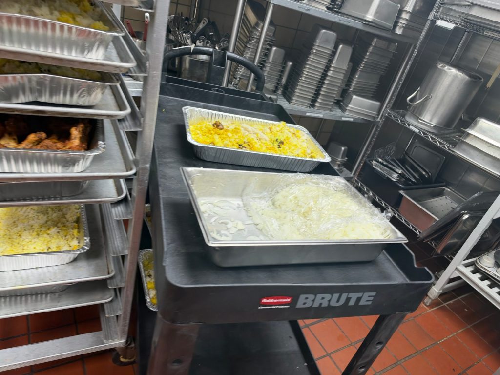 Foil pans of yellow rice and roasted chicken on a rolling rack and kitchen cart, beside a tray of sliced onions.