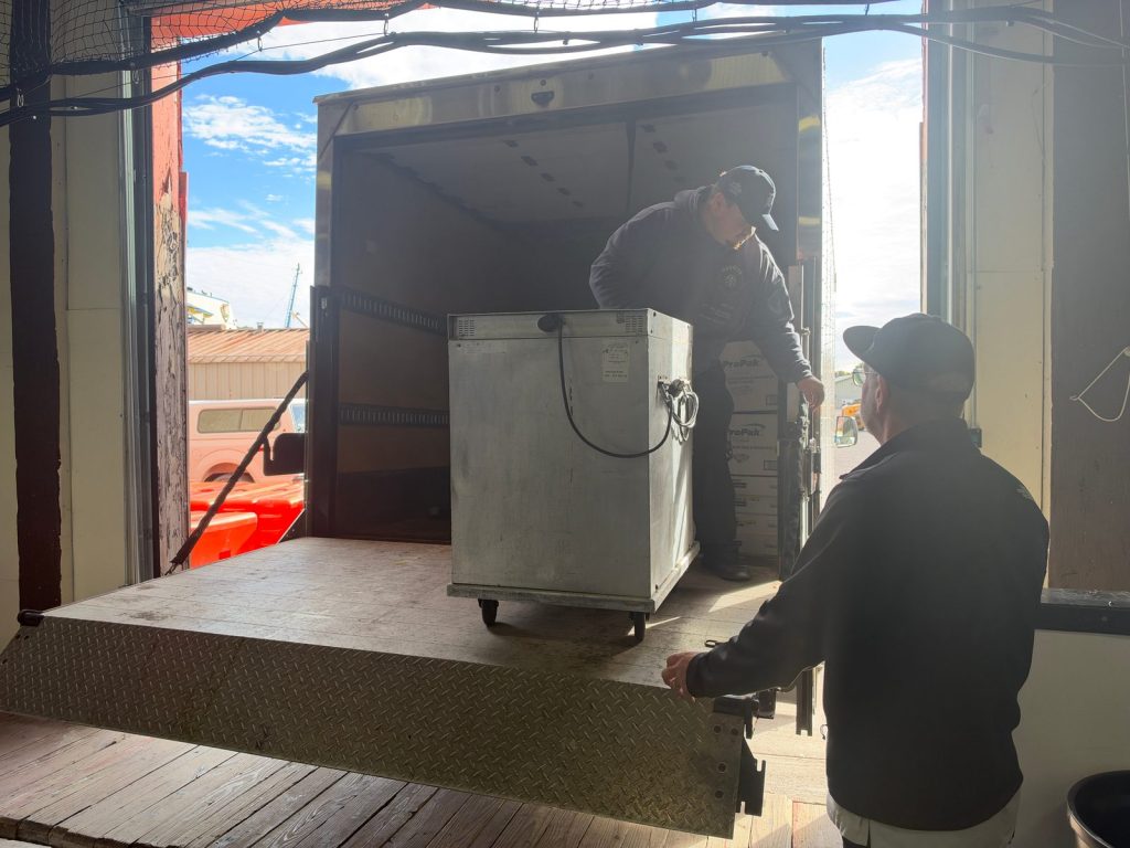 Two men move a metal food cabinet on the lift gate of a box truck.