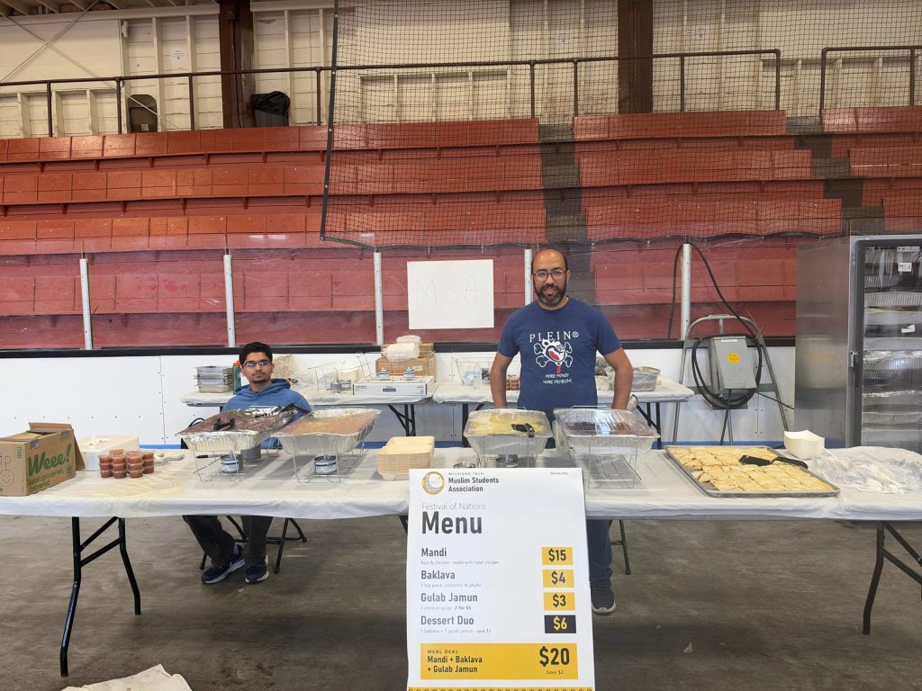 Two volunteers behind the MSA table, set with chafing dishes, baklava and a Festival of Nations menu sign.