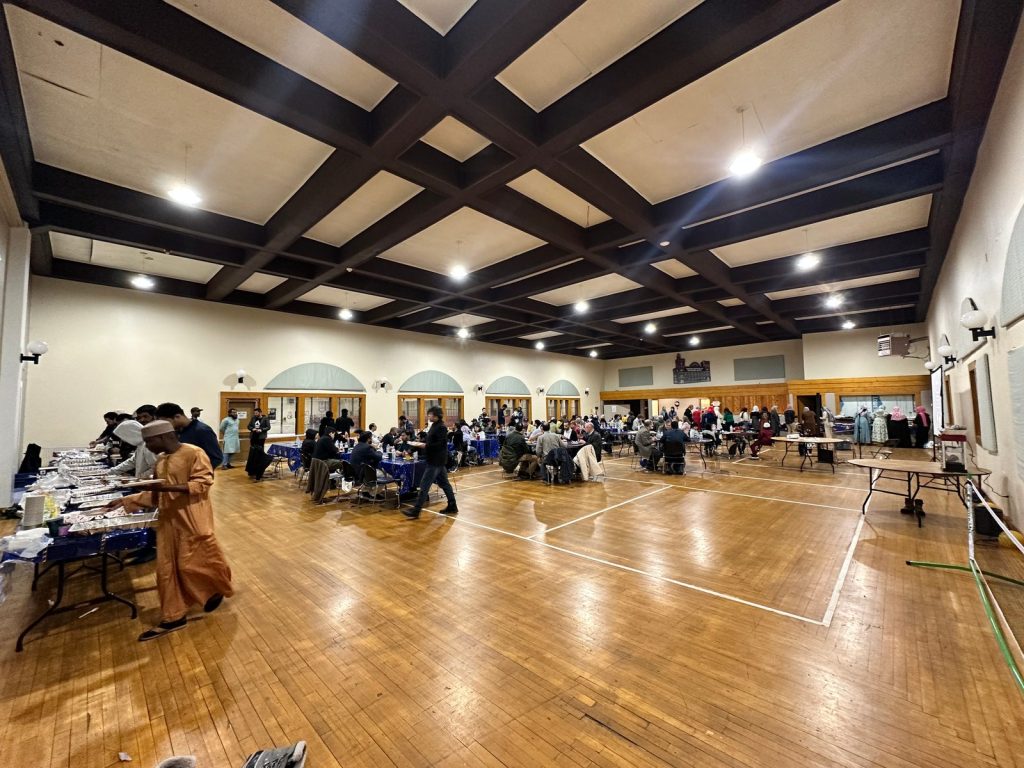 A wide view of the ballroom as guests fill plates at a long buffet and the tables fill up behind.