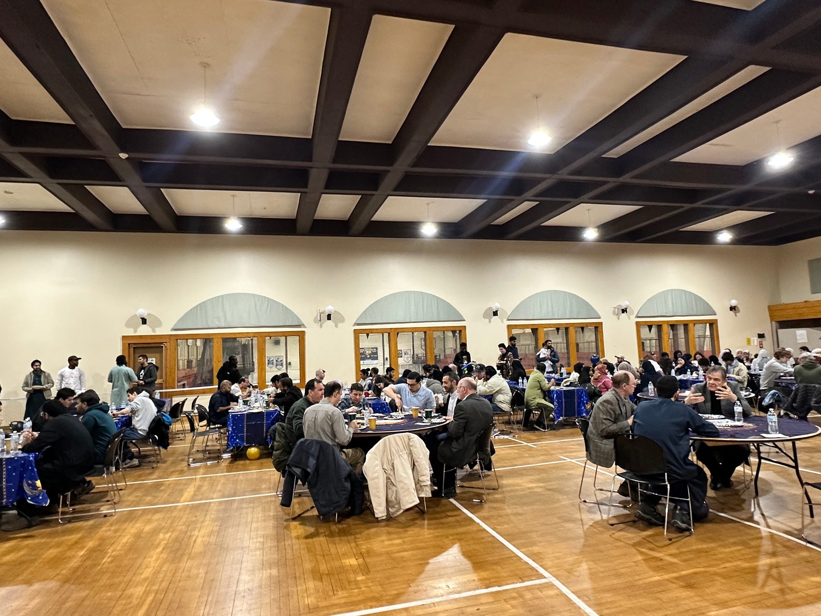 Guests eat and talk at round tables with blue tablecloths across the Dee Stadium Ballroom, under a dark beamed ceiling.