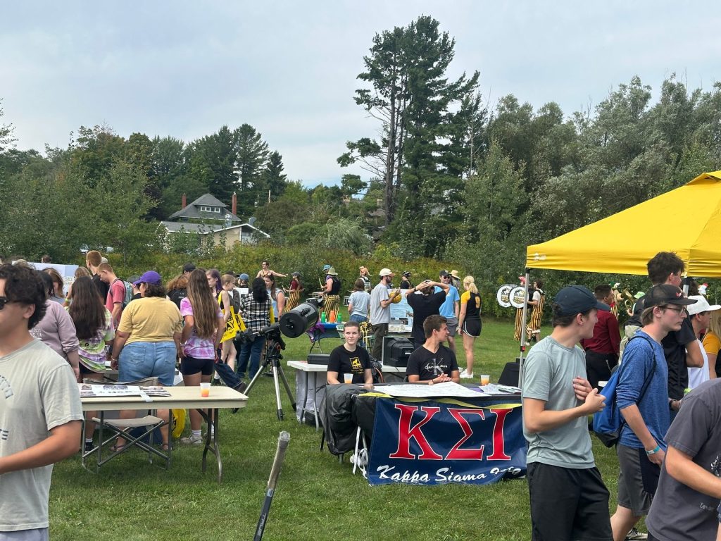 Students crowd between club tables on the lawn as a pep band in striped overalls plays in the background.