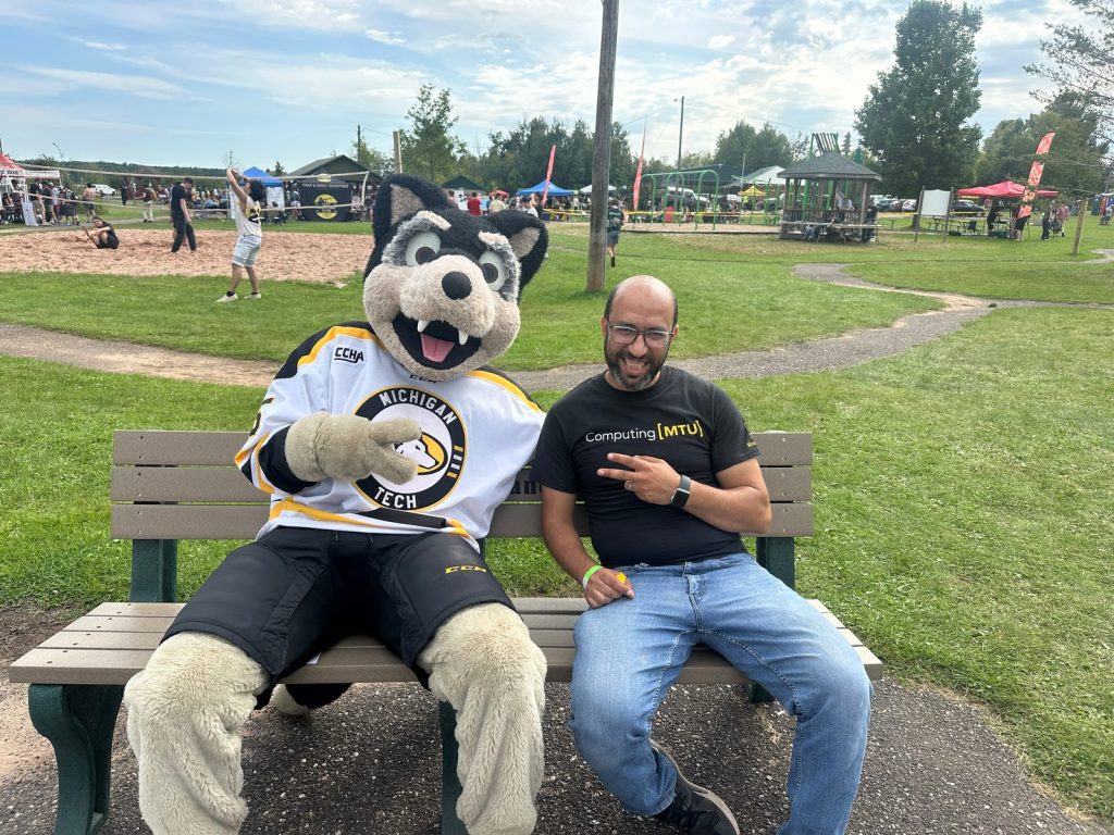Michigan Tech's husky mascot sits on a park bench beside a smiling man, with a sand volleyball court behind.