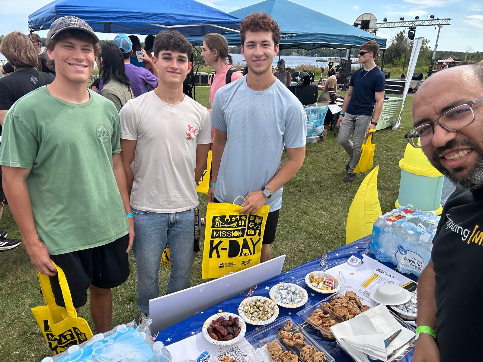 Three visitors holding yellow K-Day tote bags smile at the MSA snack table, with tents and a lake behind.