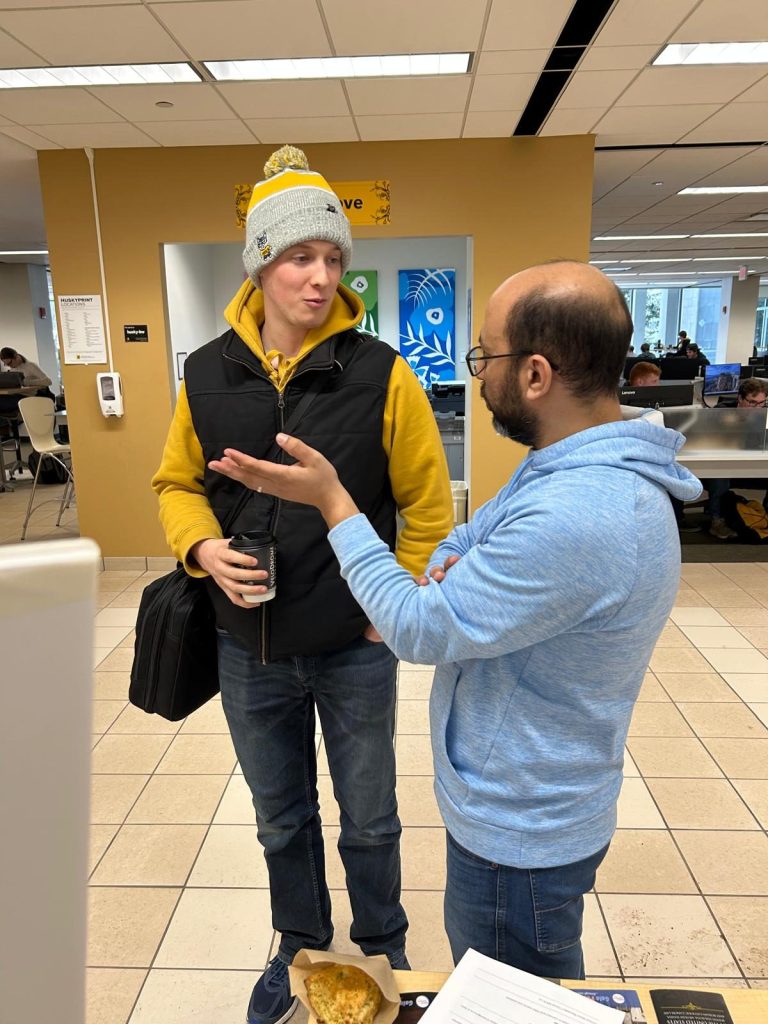 A man in a light blue hoodie talks with a visitor in a gray-and-yellow beanie holding a coffee cup.