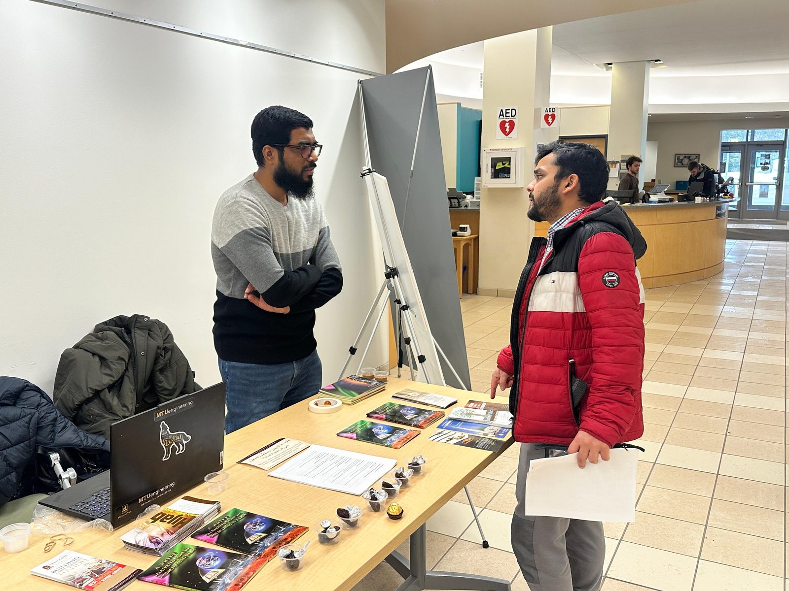 A man in a gray-and-black sweater talks with a visitor in a red jacket across a table of pamphlets in the library.
